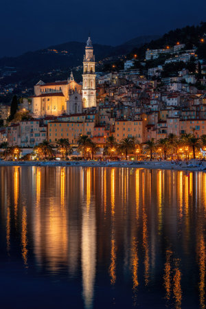 A nighttime view showcases a coastal city with brightly lit buildings, including a prominent tower. The lights reflect beautifully in calm water. The scene is likely outdoors, featuring a cool palette with warm highlights from the artificial lighting. This image is suitable for various commercial uses, such as illustrating urban environments or tourism.の素材