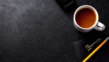 A white coffee cup sits on a dark, textured surface, alongside office supplies. The image displays a high-angle composition with a focus on contrasting colors and textures. It may be suitable for websites, editorial content, and various design projects, emphasizing a workspace setting.の素材