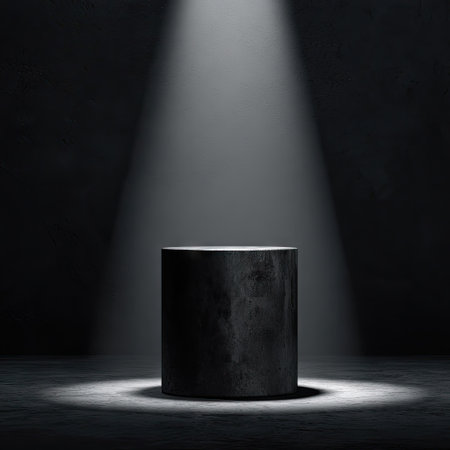A dark cylindrical pedestal is centrally positioned, illuminated by a focused overhead spotlight. The composition features a textured, dark background creating a dramatic contrast. The lighting highlights the form, suggesting a stage or product display setting. This versatile image suits various commercial applications, including product showcasing.の素材