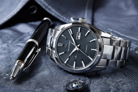 A close-up captures a luxury wristwatch alongside a pen resting on a textured fabric. The metallic watch features a dark dial, while the pen is black and silver. The scene uses soft lighting, highlighting textures and reflecting a sophisticated design. This arrangement would suit commercial applications, emphasizing style and timekeeping.の素材