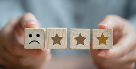 A close-up captures hands arranging wooden blocks displaying a customer satisfaction evaluation. The blocks showcase star symbols and a sad face, conveying a rating system. The composition uses neutral tones, possibly indoors under soft, diffused lighting. Suitable for articles on service reviews or consumer behavior in general.の素材