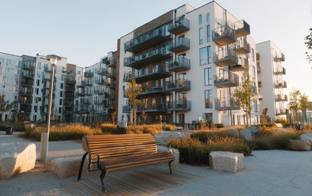 The image features contemporary apartment buildings with multiple stories, balconies, and large windows. A wooden bench sits in front of the buildings. The scene is illuminated by daylight with a warm tone, suggesting an outdoor setting. This image could be used for architectural concepts or property marketing.の素材