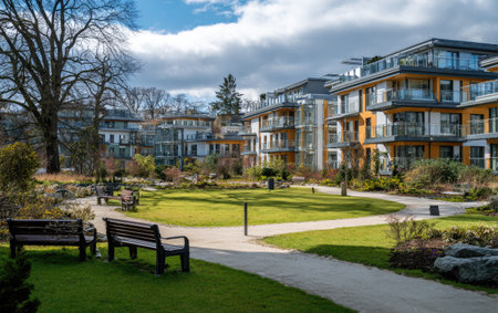 An outdoor scene showcases contemporary apartment buildings with multiple stories and balconies. The image features vibrant green grass, walkways, and manicured gardens. The setting appears to be a residential complex, with sunlight and a blue sky with clouds. The image could be utilized in commercial projects or editorial content.の素材