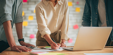 A group of individuals collaborates at a wooden table, possibly in an office environment. The scene involves a laptop, documents, and individuals taking notes. The composition is well-lit, suggesting a modern or creative workspace. This image could be suitable for illustrating teamwork or business concepts.の素材