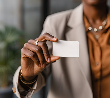 A person in a neutral-toned blazer holds a blank card. The image shows a close-up of a hand with a ring, set against a blurred background. The lighting is soft and natural. This image could be useful for business presentations, marketing materials, and other promotional applications.の素材