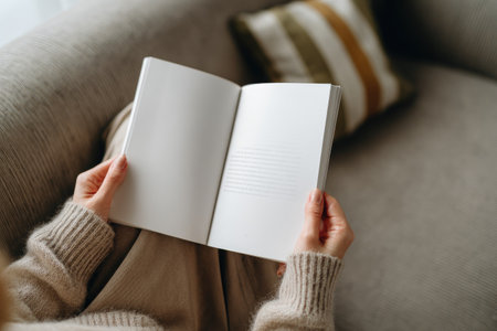 An individual holds an open book, its blank pages facing the viewer. The person is seated, possibly indoors, on a sofa with a textured fabric. Warm tones dominate the scene, with the presence of natural lighting. Suitable for illustrating ideas related to reading, education, or creative writing.の素材