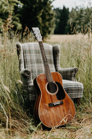 An acoustic guitar rests against a vintage armchair in an outdoor setting. The image displays a wooden guitar with a dark soundhole and fretboard, complemented by the neutral tones of the chair. The composition, bathed in natural light, suggests a serene atmosphere suitable for a variety of media applications.の素材