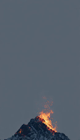 An image captures a mountain peak, seemingly ablaze with fire, contrasted against a muted gray sky. The visual texture shows the rough surface of the mountain. The composition creates a dramatic contrast in an outdoor, nighttime setting. It would be suitable for various commercial uses, including artistic designs and editorial content.の素材