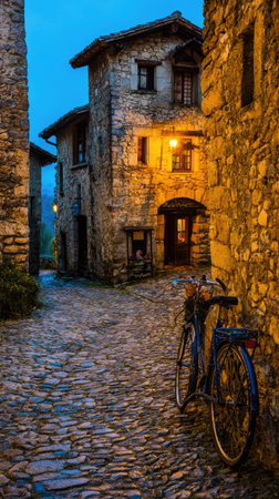 An evening scene depicts a narrow, cobblestone street flanked by aged stone buildings. A vintage bicycle rests against a building, illuminated by warm artificial light. The composition features a vertical format with a moody atmosphere. Suitable for use in a variety of commercial and editorial contexts.の素材