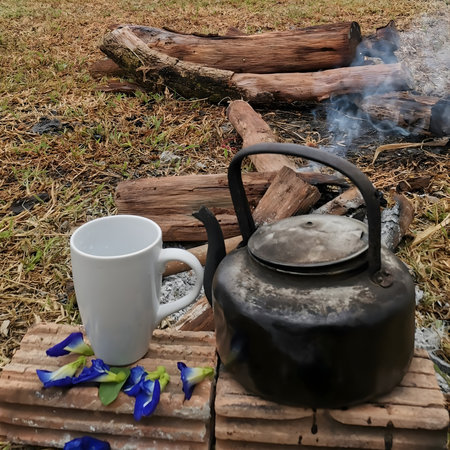 A rustic black stovetop kettle resting on brick blocks beside a simple white mug. The scene captures a natural, cozy outdoor atmosphere with a touch of vintage charm.の写真素材
