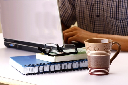 Coffee mug, stack of books and man working on a laptop computer in the backgroundの写真素材