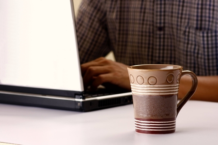 Coffee mug and man working on a laptop computer in the backgroundの写真素材