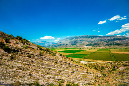 Tilted sedimentary strata descending towards a mosaic of irrigated agricultural fields set against the NemÃ«rÃ§ka mountain massif beneath an azure skyの写真素材