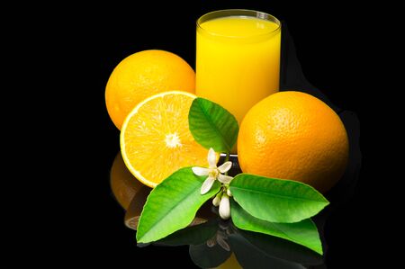 Oranges with green leaves and flowers. Orange juice in a glass cup. Still life with citrus fruit on a black background and with a dark reflection.の写真素材