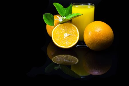 Oranges with green leaves and flowers. Orange juice in a glass cup. Still life with citrus fruit on a black background and with a dark reflection.の写真素材