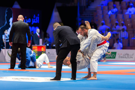ABU DHABI, UAE - APRIL 19, 2016: ABU DHABI WORLD PROFESSIONAL JIU-JITSU CHAMPIONSHIP 2016 in the IPIC ARENA. Men in white kimonos fighting on the mat. Referee next to them.のeditorial素材