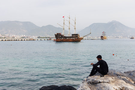 A guy sits on a rock near the sea. In the background there is an old pirate ship.の写真素材