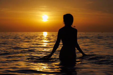 Silhouette of a girl in the sea looks at you at sunset.の写真素材