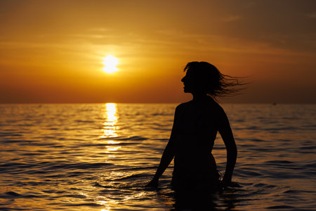 Silhouette of a girl with flying hair at sunset stands in the sea.の写真素材