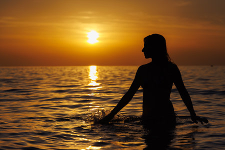 Silhouette of a girl looking at the sun and touching the sea at sunset.の写真素材