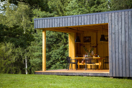 Modern cabin in a tranquil forest setting. Contemporary design blends with nature. Peaceful retreat with a large tree and outdoor seating. Perfect for architecture, travel, or nature projects.の写真素材