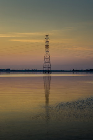 Calm lake or river at sunset with a silhouette of trees and an electrical tower. Beautiful sky with warm colors reflected in the water. Peaceful and serene landscape.の写真素材