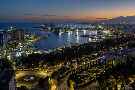 Malaga port shows boats and lights as dusk falls. People walk along the waterfront where trees line...