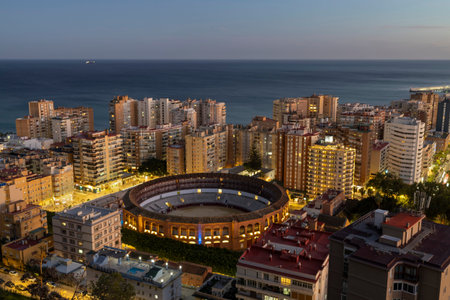 Malaga, Spain at sunset. The bullring is highlighted among buildings. The coastline is visible in...