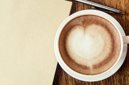 A cup of coffee with heart shape, pen and blank book on wood backgroundの写真素材