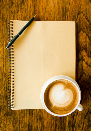 A cup of coffee with heart shape, pen and blank book on wood backgroundの写真素材