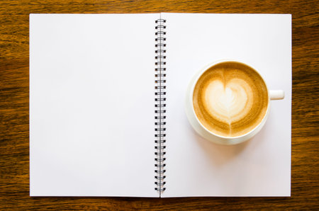 A cup of coffee with heart shape and open blank book on wood backgroundの写真素材