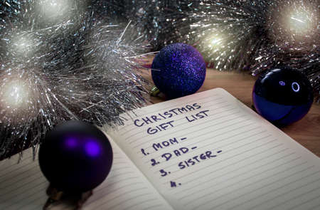 Blue Christmas baubles, a notepad and dim lights. An English text handwritten in a paper notebook "Christmas gift list - mom, dad, sister".の写真素材