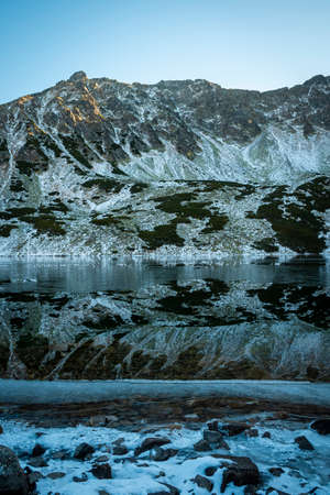 Shadowy valley in Tatra Mountains. Winter is coming, the lake is freezing, stillness in the water makes a perfect mountain ridge reflection. Selective focus on the rocks, blurred background.の写真素材