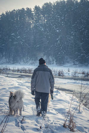 Man with a dog walking on snow, Neris Regional Park, Lithuania. Cold January day hike on a frozen riverside. Selective focus on the animal, blurred background.の写真素材