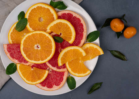 Sliced citrus fruits, grapefruits, lemons, oranges, with green leaves on a white plate. Top view with visible textureの写真素材