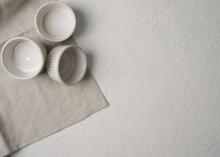 Flat lay of three empty white ceramic ramekins stacked against white background, served linen napkin with copy space. Top view ramekin, prepare for cooking. Selective focus.の写真素材