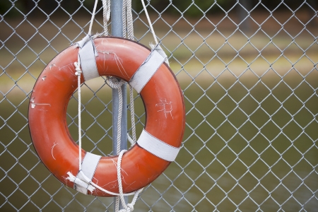 A rugged orange life ring hangs on a chain link fence outside in the summer の写真素材
