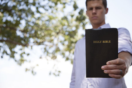 A young, confident man stands outside holding a black bible with one hand.