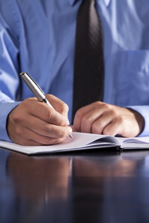 A young businessman writes in a notebook while sitting at a desk.の写真素材