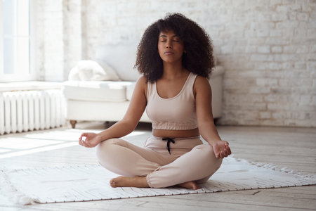 A black woman sits in a lotus position. Place hands on lap, fingers folded, eyes closed, and meditate. Doing yoga at home. Anxiety reduction concept, no stress, healthy habit, mindfulness lifestyle.の写真素材