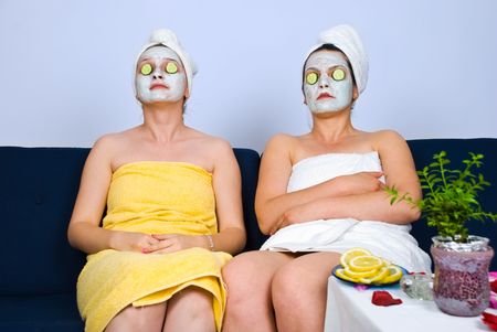 Two women sitting on sofa at spa retreat and having facial masks on face and slices cucumber on eyesの写真素材