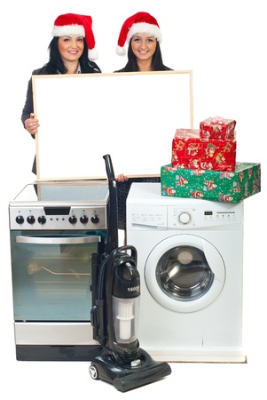 Two beautiful women with Santa hats holding blank banner and making Christmas offer at household appliancesの写真素材