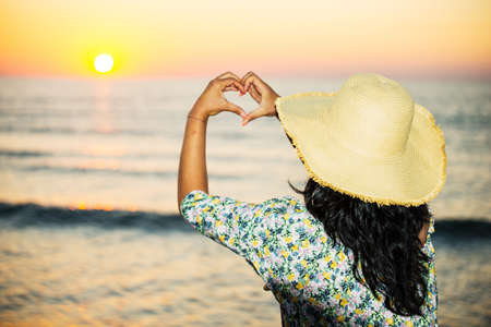 Back of woman with hat makes a heart out of her hands to the sunrise at seaの写真素材