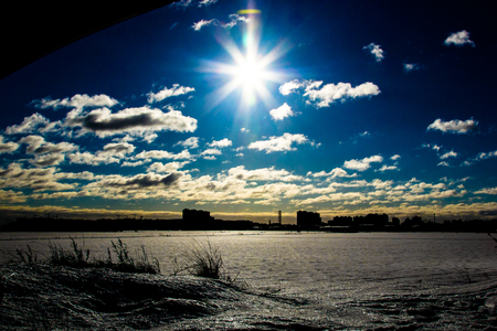 Winter landscape, picturesque view of high-rise buildings of one of the districts of the city, blue sky with white clouds in the rays of light, bright sun over the snowy plain with dried grass on the front, the silhouette of a distant cityの写真素材