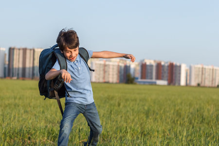 The schoolboy happily runs with a backpack on the field against the background of the city landscape