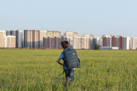 The schoolboy happily runs with a backpack on the field against the background of the city landscape