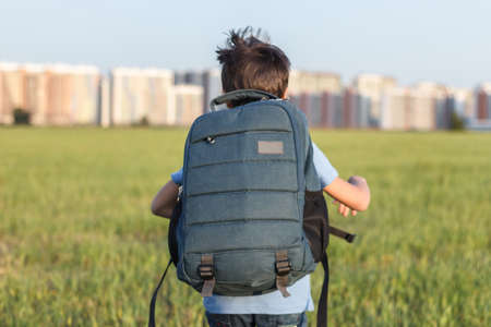 The schoolboy happily runs with a backpack on the field against the background of the city landscape.の写真素材
