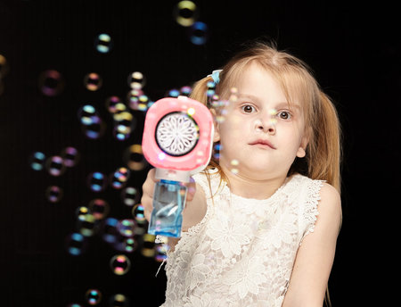 Child blonde girl 6 years old in a white dress on a dark background among air bubbles