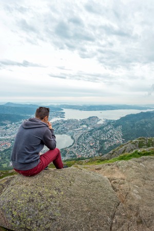 Young man in sportswear is sitting on cliff's edge and looking to misty valley bellow, Bergen, Norwayの写真素材