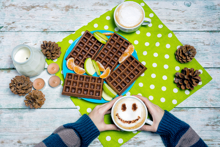 Cappuccino coffee with smiley face with waffles on wooden table in female hands overhead viewの写真素材